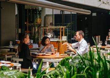 Attain on 5th apartment homes with Three people sit at an outdoor restaurant table, engaged in conversation, surrounded by green plants.